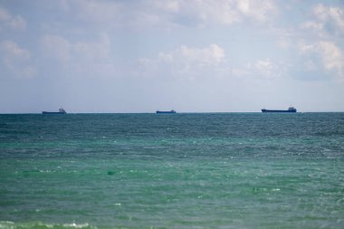 Sakin yeşil-mavi kıyı suları tarafından çerçevelenmiş yumuşak bulutlu bir gökyüzünün altında, uzak ufukta sürüklenen üç kargo gemisi..