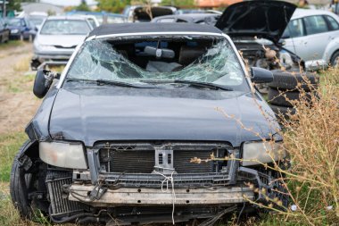Parçalanmış ön camı ve açık kaputu olan hasarlı siyah araba diğer hurdalıkların arasında terk edilmiş bir hurdalıkta duruyor..