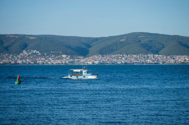 Bulgaristan 'ın arka planında bir dağ kasabası ve ormanlı dağlarla birlikte sakin kıyı sularındaki bir şamandıranın yanından kayan beyaz bir tekne görülüyor.
