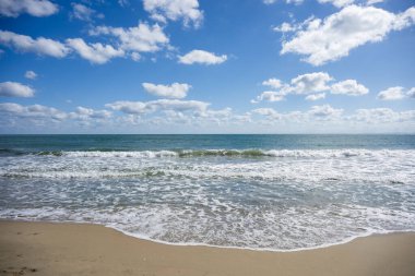 Yumuşak dalgalar, yumuşak bulutlar ve açık bir ufuk ile huzurlu sahil sahnesi.