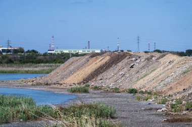  Enkaz ve toprak yığını, arka planda sanayi binaları ve bacaların yükseldiği sakin bir su yolu ile sınırdır..