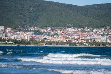 Yoğun inşa edilmiş bir sahil kenti, açık bir gökyüzünün altındaki yeşil bir yamaca karşı yükselirken, dalgalar kıyıya doğru kıvrılıyor..