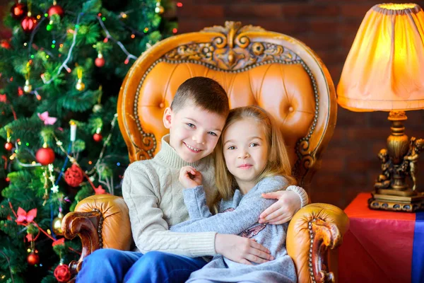 brother nestlessly hugs his sister sitting in a chair near the holiday tree, smiling together