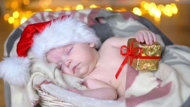 サンタ クロースの帽子で生まれたばかりの赤ちゃんバスケットの中にある 小さなギフト ボックスを保持 — ストック動画