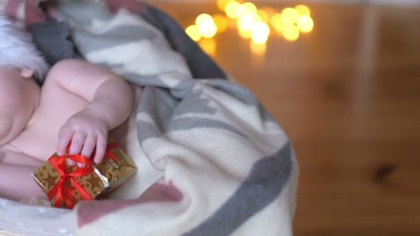 クリスマスの帽子は 明るいお祭りの背景にささやかな贈り物を彼の手を保持しているバスケットに横たわって眠っているサンタ クロースの生まれたばかりの赤ちゃん — ストック動画