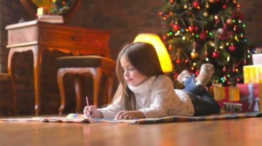 Beyaz örgü kazak güzel küçük kız yerde yatıyor ve şenlikli bir Noel ağacı ve bir sürü hediye Noel Baba'ya bir mektup arka planda yazıyor