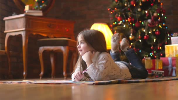 一个穿着白色针织毛衣的漂亮小女孩躺在地板上 写了一封信给圣诞老人 背景是一棵喜庆的圣诞树和一堆礼物 — 图库视频影像