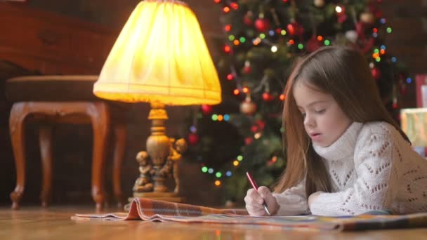 小女孩躺在房间的地板上给圣诞老人写信 在背景圣诞树上 — 图库视频影像