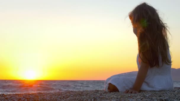 美しい夕日にビーチに座って砂と遊ぶ美しい女の子 — ストック動画