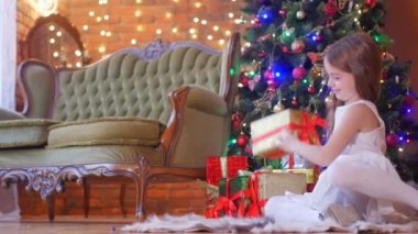 Beyaz elbiseli güzel bir kız Noel ağacının yanında yerde oturuyor kurdeleyi çözüyor ve hediyeyi açıyor, neşeli bir ruh hali.