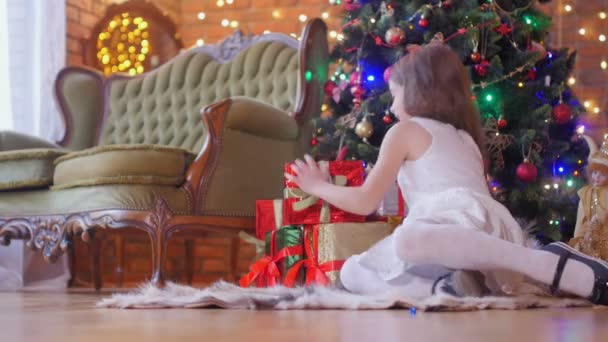一个穿着白色连衣裙的漂亮小女孩坐在圣诞树旁的地板上 解开带子 打开礼物 一种节日的气氛 — 图库视频影像