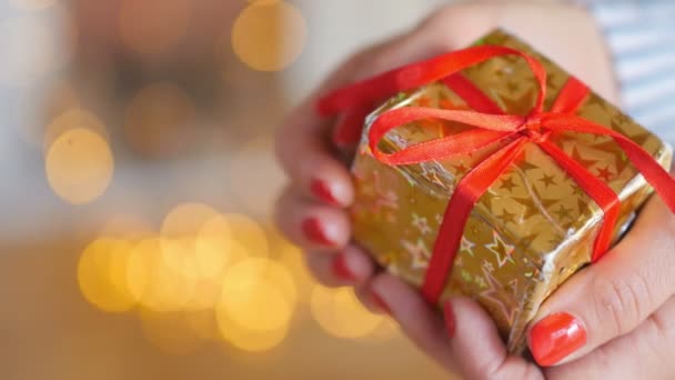 休日少女の手のクローズアップは 明るいお祝いの背景に赤いリボンで黄金の紙に包まれた贈り物と小さな箱を慎重に保持します — ストック動画