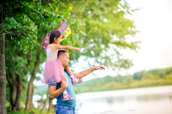 dad and little daughter are walking in the spring park, standing on the shore of the lake and something is being considered, a girl with a kite in her hands