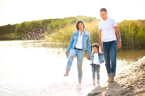 A happy family walks along the bank of the river in the evening, barefoot in the water and splashing, healthy outdoor recreation