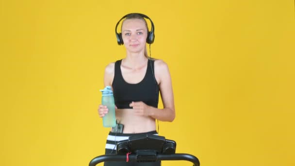 穿着运动服 头戴耳机的金发女孩在跑步机上锻炼 喝运动瓶装水 — 图库视频影像