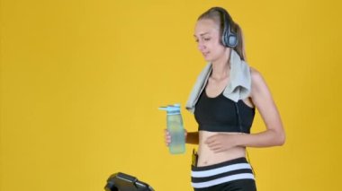 Spor kıyafetli, kulaklıklı sarışın kız koşu bandında çalışıyor ve spor şişesinden su içiyor..