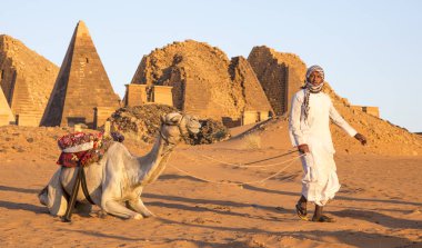 Meroe, Sudan - 19th December, 2015: Sudanese man with his camel in desert near Meroe pyramids clipart