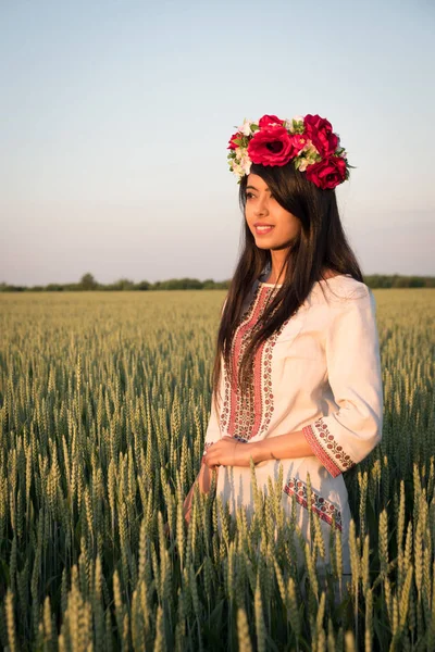 Indian woman in Ukrainian embroidery posing in fieldBeautiful indian young woman in traditional Ukrainian embroidery clothes and floral handmade wreath stand in field, look in distance dreaming or thinking, pretty ethnic woman posing in nature