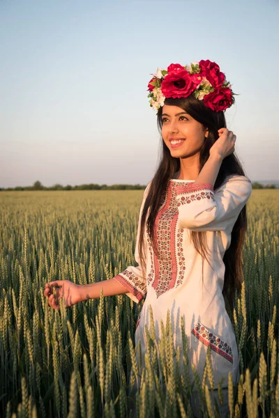 Indian woman in Ukrainian embroidery posing in fieldBeautiful indian young woman in traditional Ukrainian embroidery clothes and floral handmade wreath stand in field, look in distance dreaming or thinking, pretty ethnic woman posing in nature