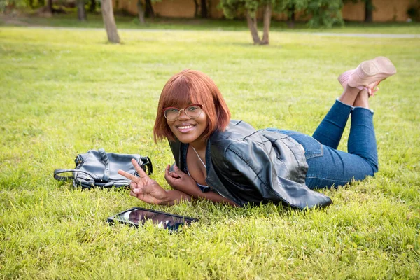 Parkta çimlerin üzerinde yatan siyah genç kız. Gülümsüyor ve kameraya bakıyor. Kız dışarıda karantinada vakit geçiriyor..