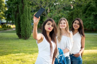 Üç etnik çeşitlilikte kız parkta selfie çekiyor. Genç ve gülümseyen kızlar. Çalışmaktan ya da işten boş zaman.