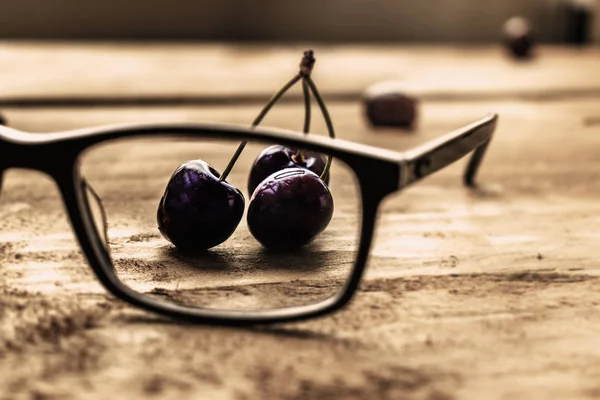 Red cherries seen through a pair of glasses, on a wooden table.