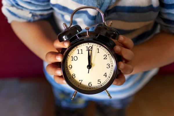 Child in pajamas holding with his hands a black alarm clock with retro style.