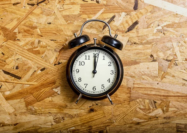 Black alarm clock marking twelve o\'clock, on OSB wood.