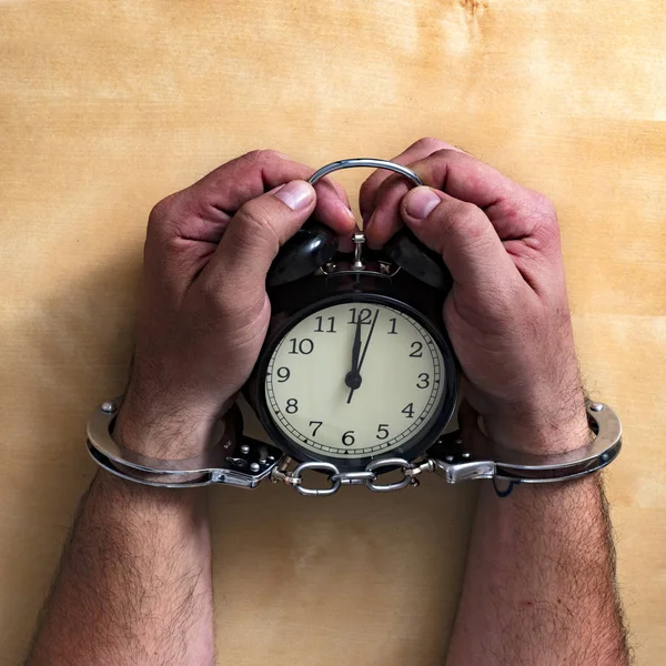 Time in jail. Shackled hands holding an alarm clock on a wooden table.