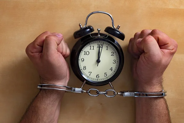 Time in jail. Shackled hands holding an alarm clock on a wooden table.