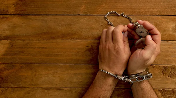 Time in jail. Chained hands holding a hand watch with chain, on a wooden table.