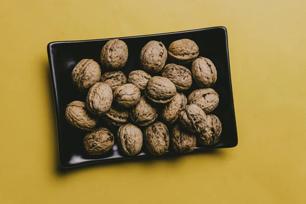 Black plain dish full of nuts on a yellow background.