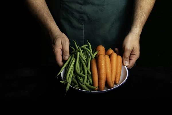 Человек в зеленом фартуке представляет овощи и овощи в саду . — стоковое фото