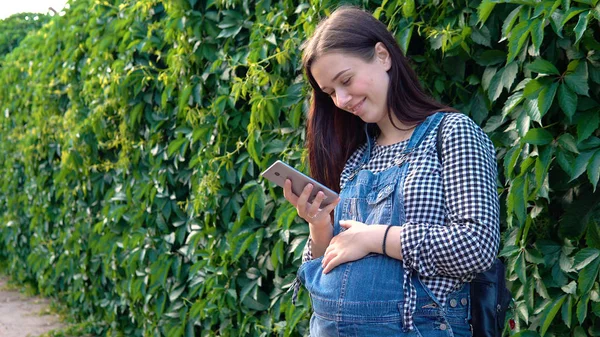 Genç hamile kız telefonu, yeşil zemin üzerine bakarak tulum giymiş.