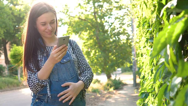 Genç hamile kız telefonu, yeşil zemin üzerine bakarak tulum giymiş.