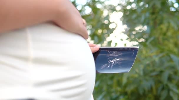 Terhes nő, ultrahanggal, fotóval vagy képpel a hasán a leendő lányának vagy fiának a karjában. A lány újszülöttet vár. Az anyaság koncepciója. Babaköszöntő. — Stock videók