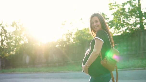 Schöne schwangere Mädchen, in einem T-Shirt und mit einem Rucksack, in den Strahlen der untergehenden Sonne, streichelt ihren Bauch und lächelt — Stockvideo