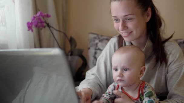 Madre joven en licencia de maternidad está trabajando desde casa, sosteniendo al bebé, sentado frente a la computadora portátil . — Vídeo de stock
