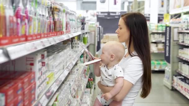 Hermosa mujer con bebé hijo de compras en el supermercado, la joven madre elige cepillos de dientes para su familia en el mercado — Vídeos de Stock