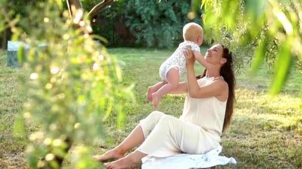 Una hermosa madre joven, en un vestido blanco con su hijo recién nacido, sentada jugando en un prado verde sobre el fondo de un olivo. Movimiento lento — Vídeos de Stock