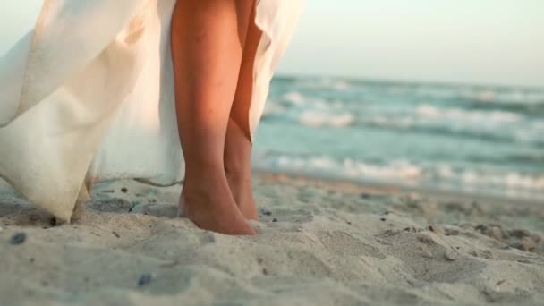 Mavi deniz kıyısında beyaz uzun elbiseli beyaz bir kızın bacakları, kum, güneş, lens üzerinde duruyor. Yavaş çekim. — Stok video