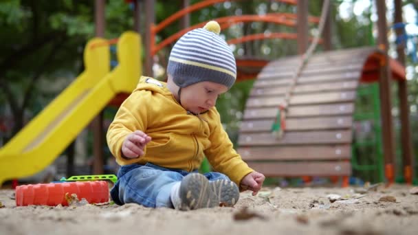 小さなかわいい男の子は、遊び場の砂の背景に座って再生されます。黄色いセーターと帽子の秋 — ストック動画