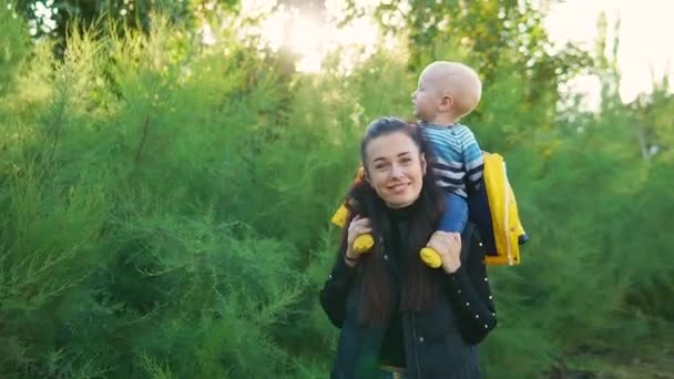 Mom carries her child in the park at sunset, in a yellow jacket, they are laughing happily. — Stock Video