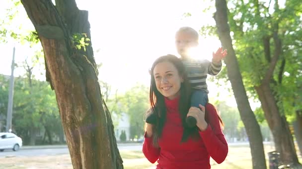 年轻的母亲和她的小儿子，微笑着，走在公园里。一个男孩坐在一个穿红色毛衣的女孩的位子上 — 图库视频影像