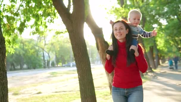 Jonge moeder en haar kleine zoon, glimlachend, wandelen in het Park. Een jongen zit schrijlings op een meisje in een rode trui — Stockvideo