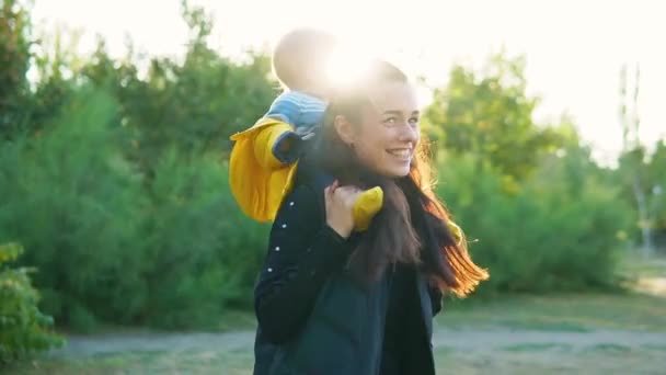 Annem gün batımında parkta çocuğuyla oynuyor. Çocuk sarı bir ceket omuzlarında oturur ve mutlu bir şekilde gülümsüyor. Birlikte dön. — Stok video