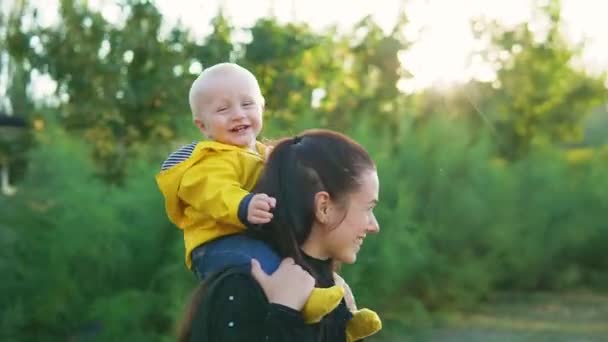 Mama gra z dzieckiem w parku o zachodzie słońca. Chłopak siedzi na ramionach w żółtej kurtce i uśmiecha się szczęśliwie. Kręć razem. — Wideo stockowe