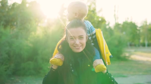 Mamá lleva a su hijo en el parque al atardecer, con una chaqueta amarilla, se ríen alegremente . — Vídeos de Stock