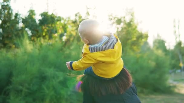 Máma hraje se svým dítětem v parku při západu slunce. Chlapec sedí na ramenou ve žlutém kabátku a šťastně se usmívá. Rotace dohromady. — Stock video