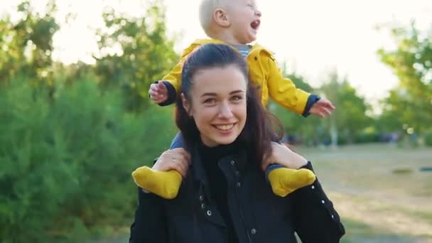 Mama gra z dzieckiem w parku o zachodzie słońca. Chłopak siedzi na ramionach w żółtej kurtce i uśmiecha się szczęśliwie. Kręć razem. — Wideo stockowe